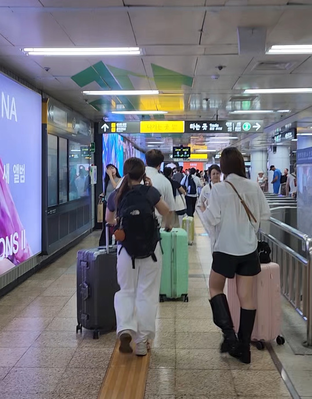 Three travellers pulling wheeled suitcases along a corridor inside Hongik Univ. Station, seen from behind.