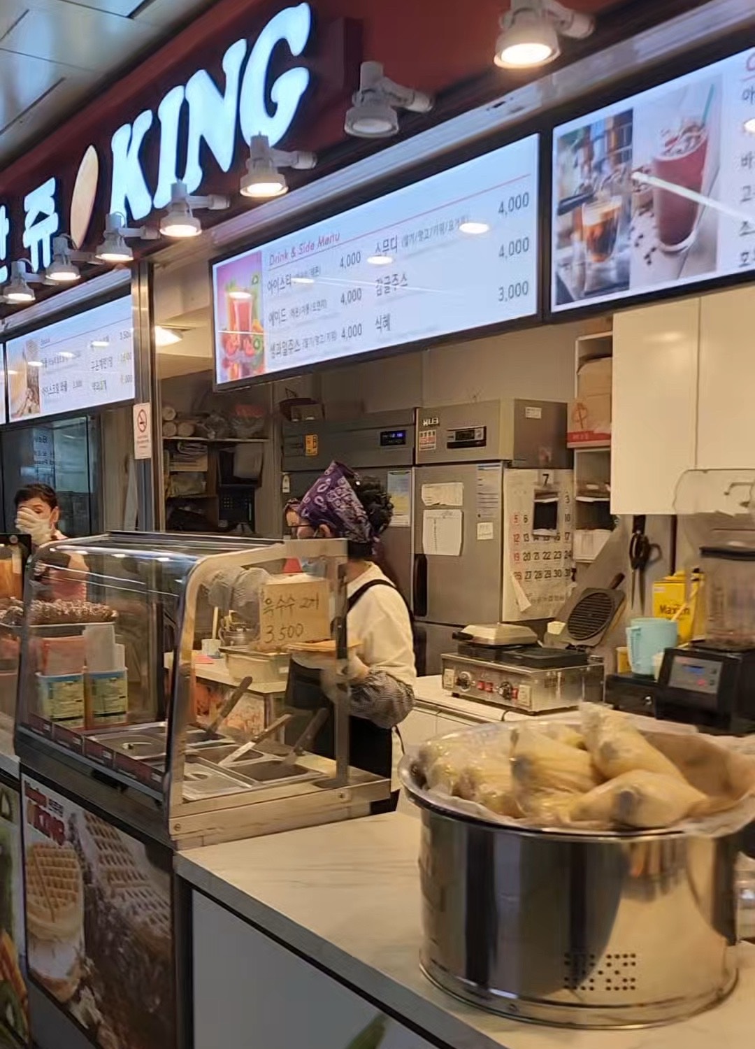A snack counter inside a Seoul subway station, with steamed corn in a large pot, a waffle iron, drink cases and a price board overhead.