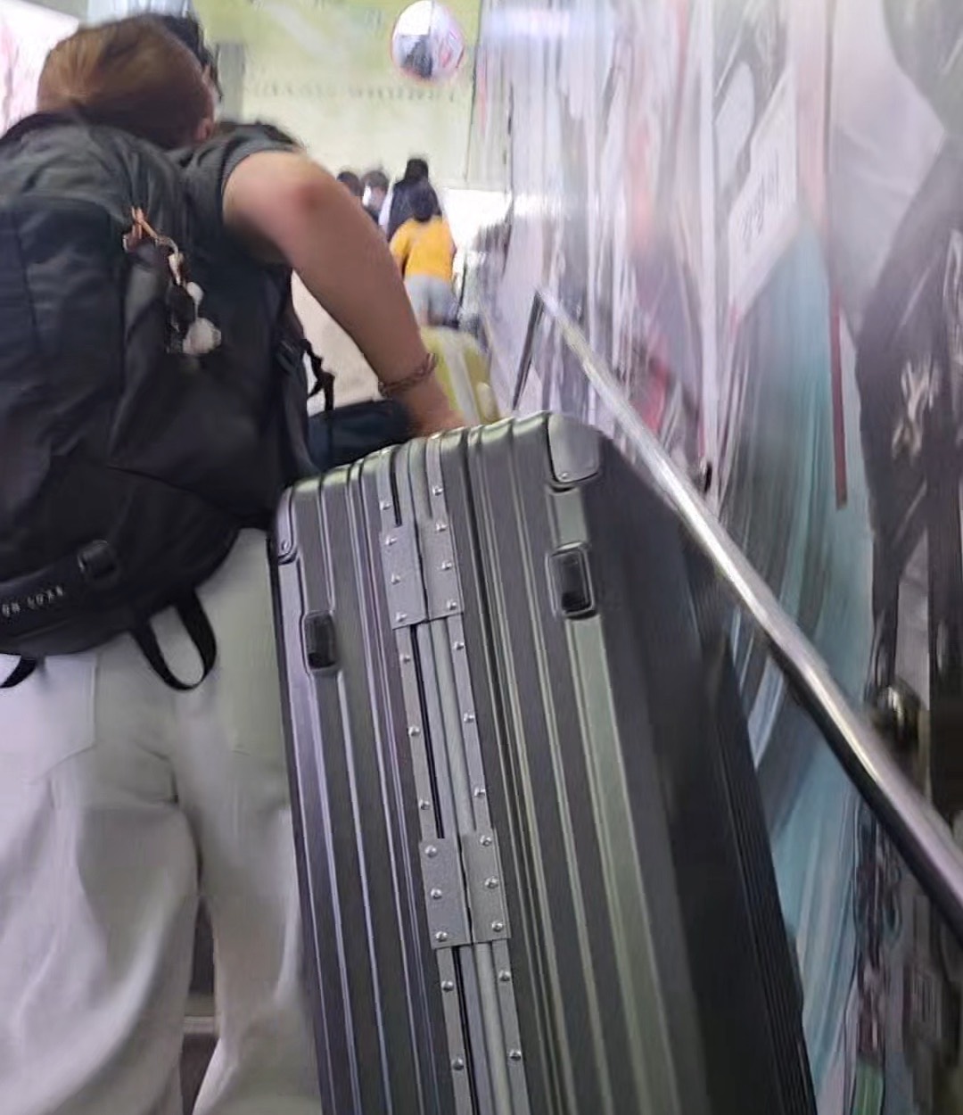 A traveller with a backpack hauling a large hard-shell suitcase up a flight of stairs in a Seoul subway station, seen from behind.